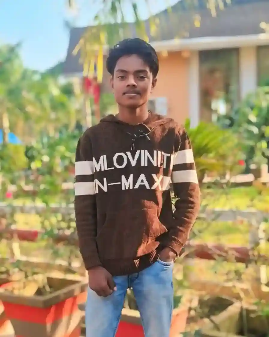 Young man wearing brown hoodie with text 'MLOVIN IT N-MAX' and blue jeans, standing outdoors in a sunny garden with greenery background"
