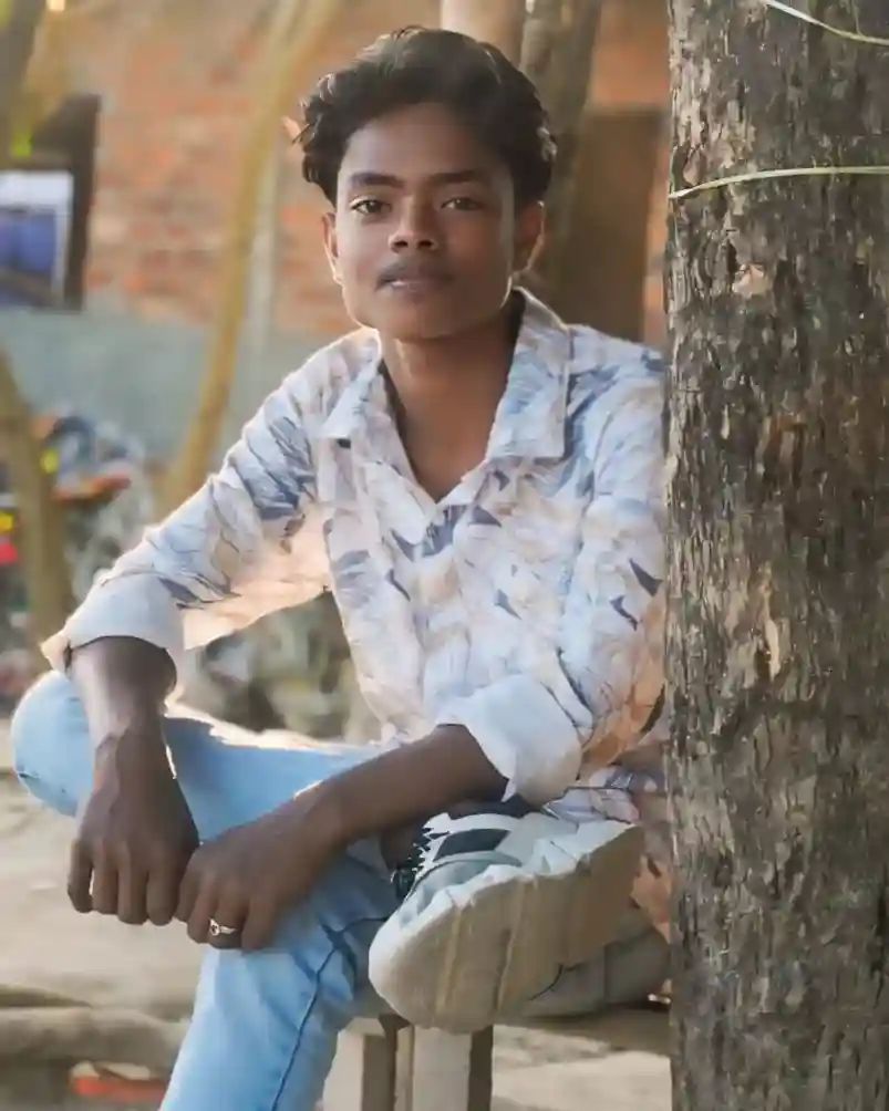 Young man wearing brown hoodie with text 'MLOVIN IT N-MAX' and blue jeans, standing outdoors in a sunny garden with greenery background"