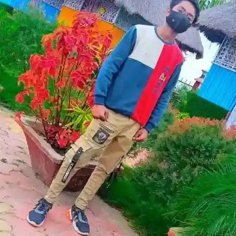 Young man wearing brown hoodie with text 'MLOVIN IT N-MAX' and blue jeans, standing outdoors in a sunny garden with greenery background"