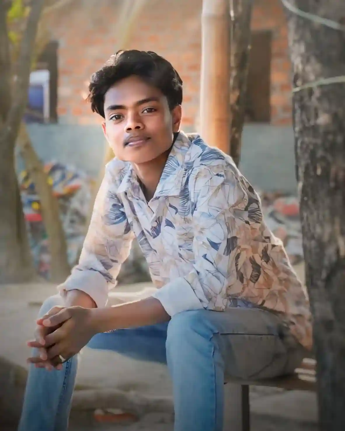 Young man wearing brown hoodie with text 'MLOVIN IT N-MAX' and blue jeans, standing outdoors in a sunny garden with greenery background"